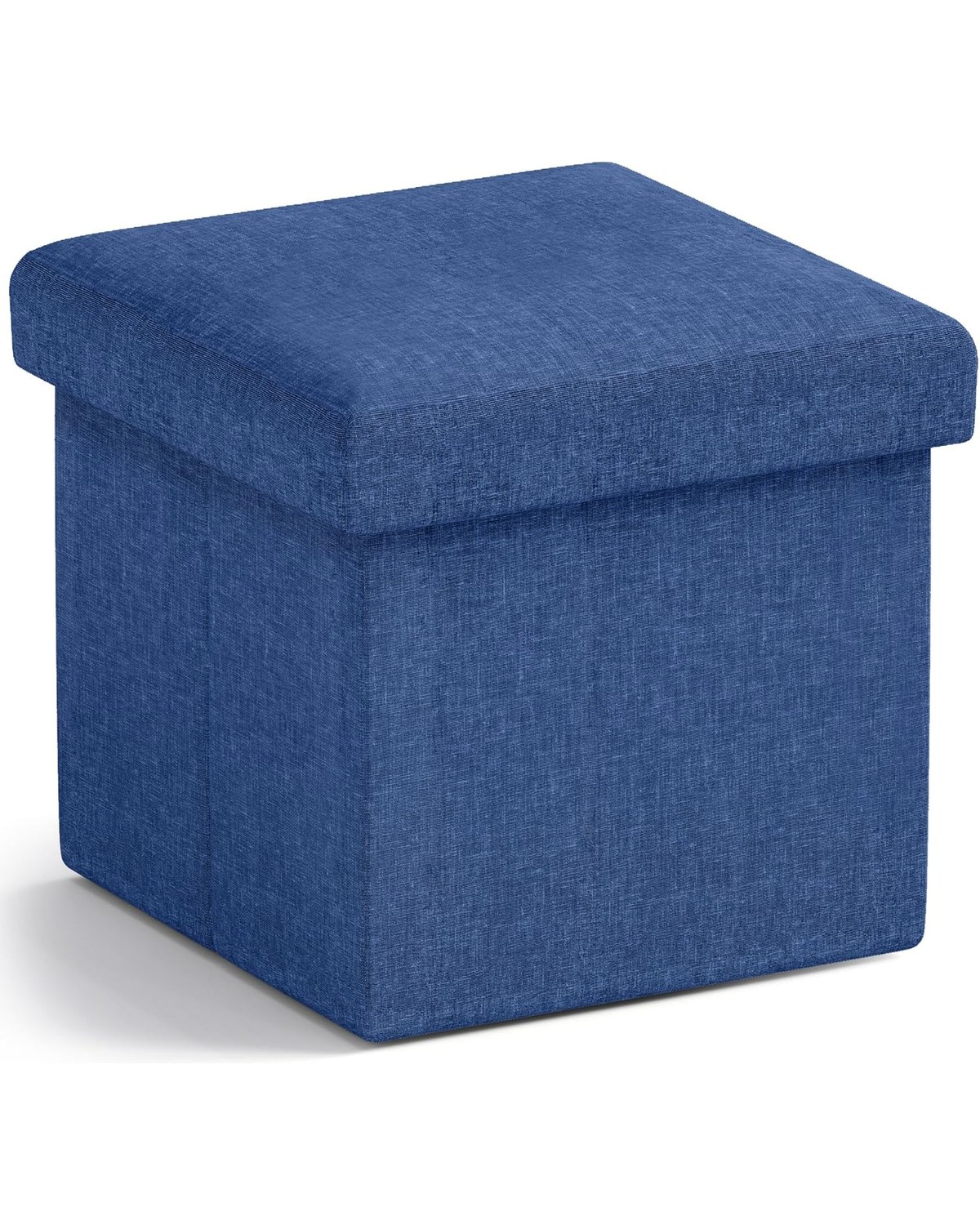 A neutral grey fabric cube ottoman in a small dorm room, lid lifted to show interior storage holding extra blankets and shoes, used as a footrest in front of a desk chair.