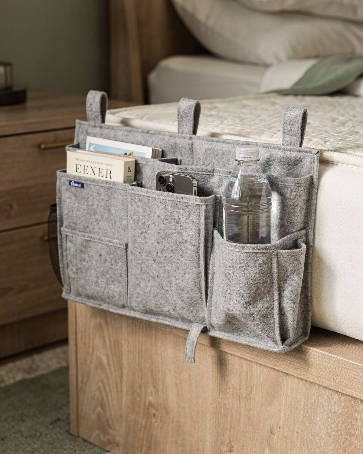 A grey felt hanging caddy with multiple pockets draped over the side of a bunk or dorm mattress, holding a phone, books, and a water bottle.