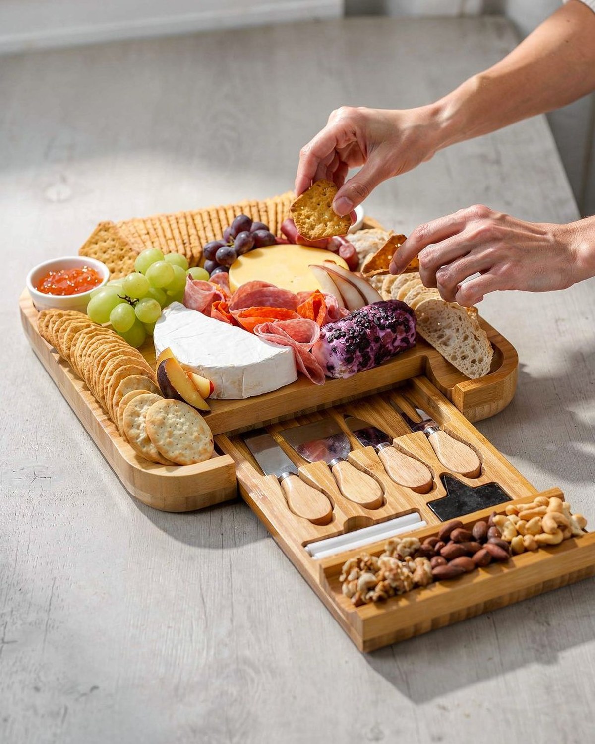 A large round bamboo charcuterie board with a hidden slide-out drawer of cheese knives, styled with crackers, fruit and cheese.