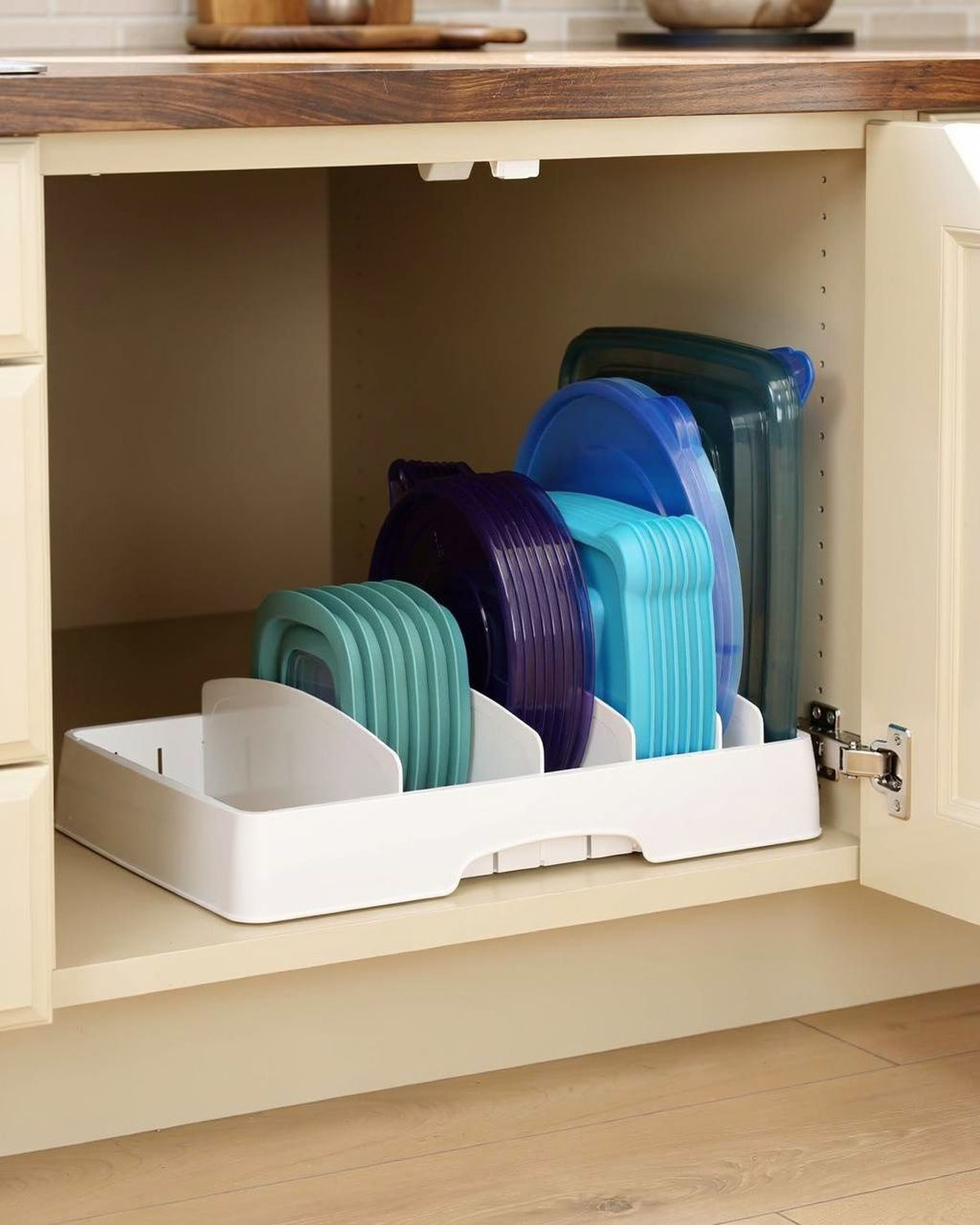 A white plastic in-cabinet caddy with five adjustable vertical dividers holding food-storage lids upright by size.