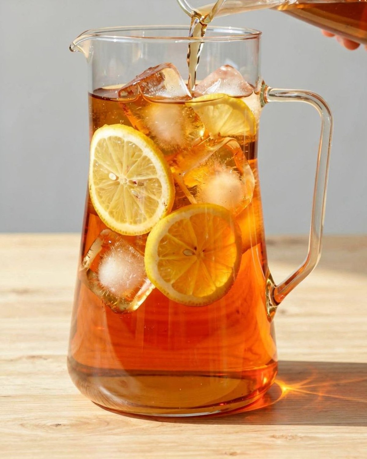 A clear borosilicate glass pitcher with a curved spout and handle, filled with iced tea and citrus.