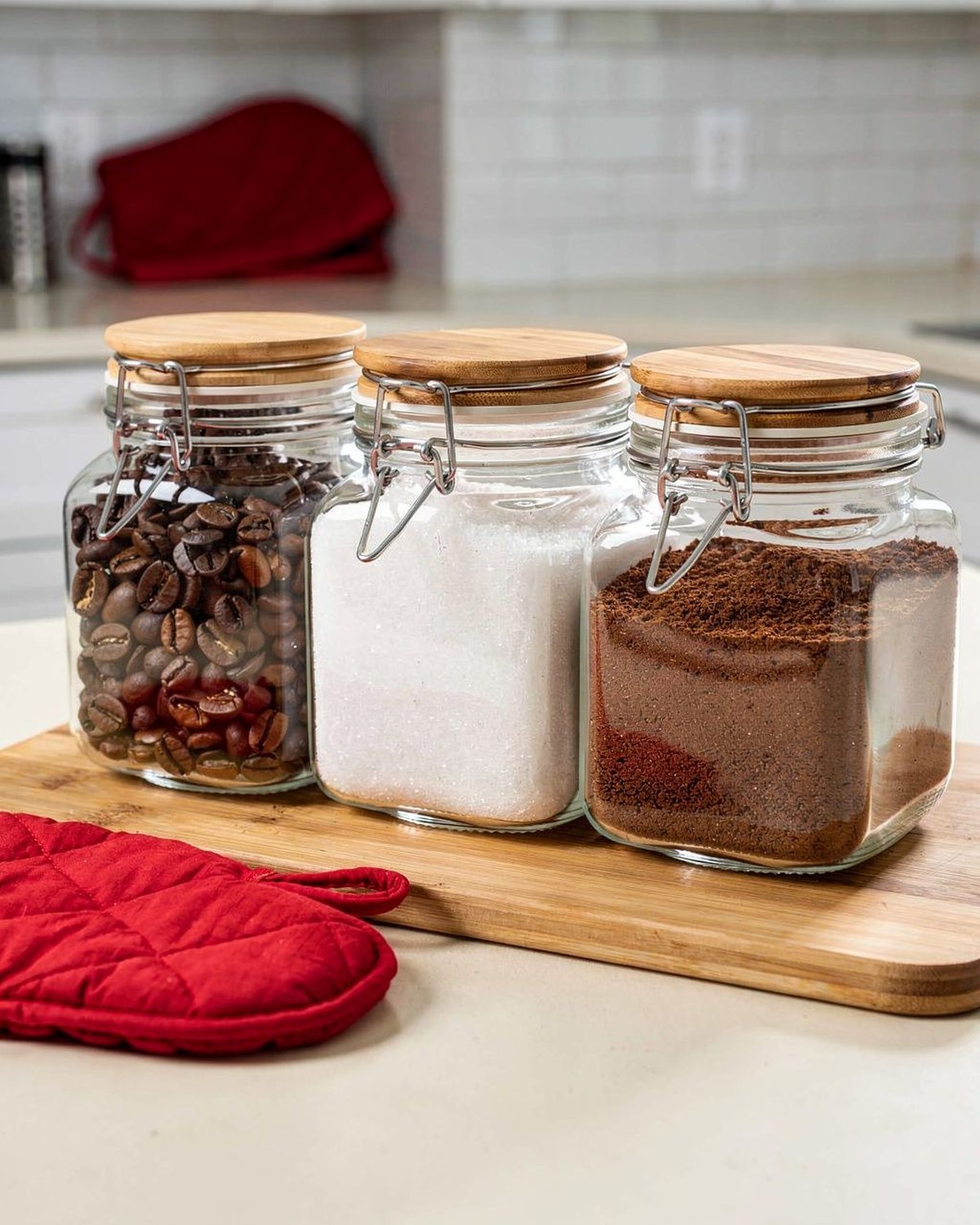Three square clear-glass jars with bamboo clamp lids holding coffee beans, sugar, and ground coffee in a row.