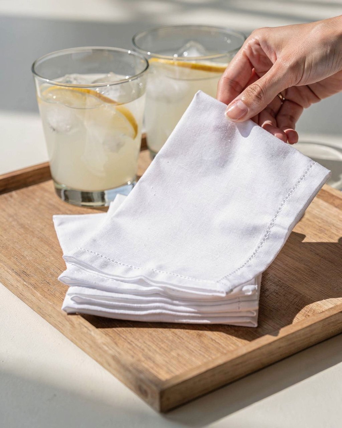 A stack of soft cotton cocktail napkins with a thin hemstitch border, fanned beside drinks on a tray.