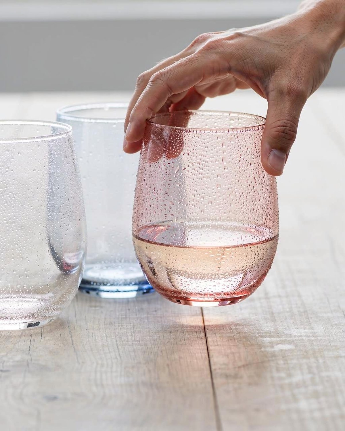 A set of clear shatterproof acrylic stemless wine glasses, beaded with condensation, on a poolside table.