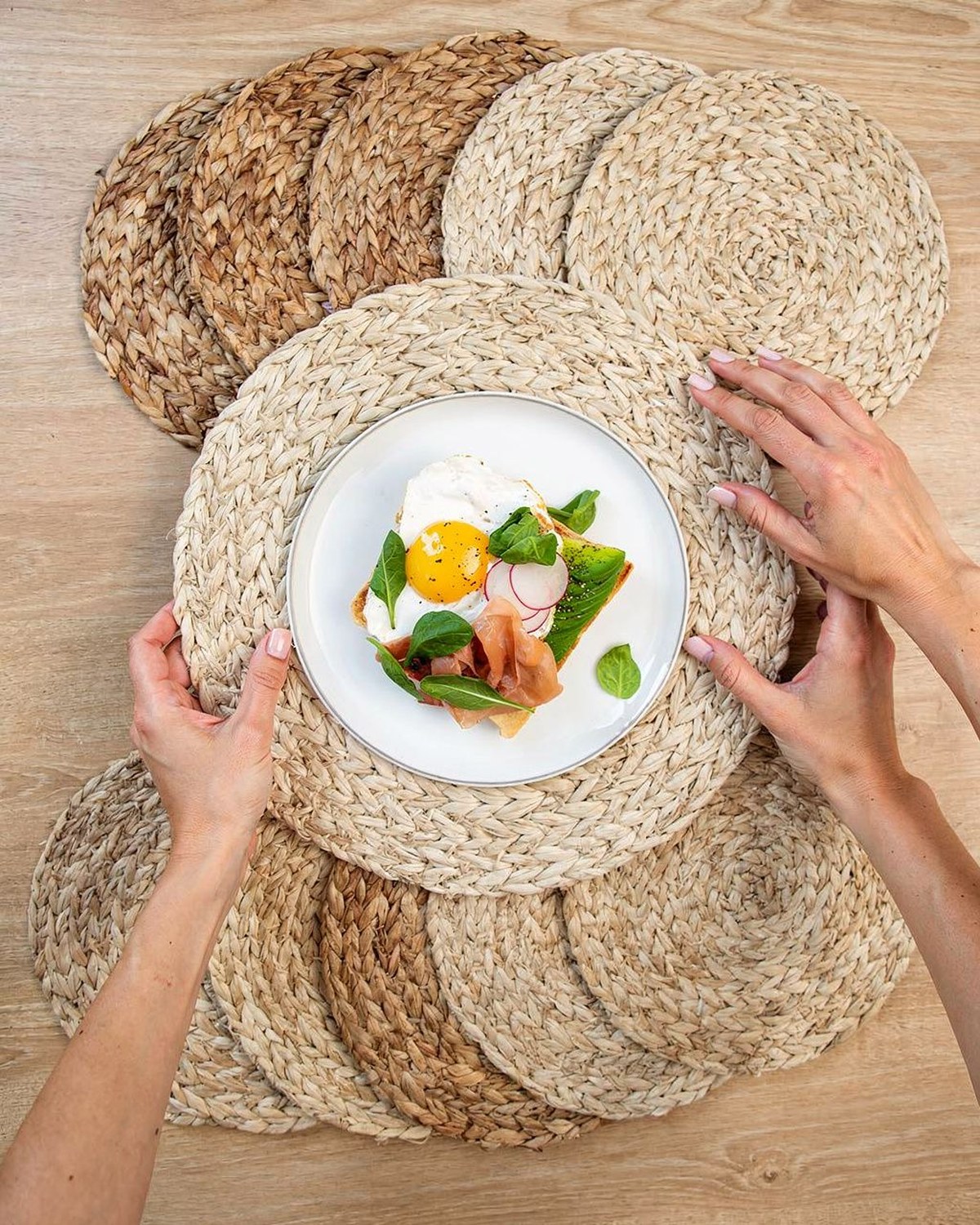 Six round natural water-hyacinth woven placemats stacked and fanned, with one set under a summer place setting.