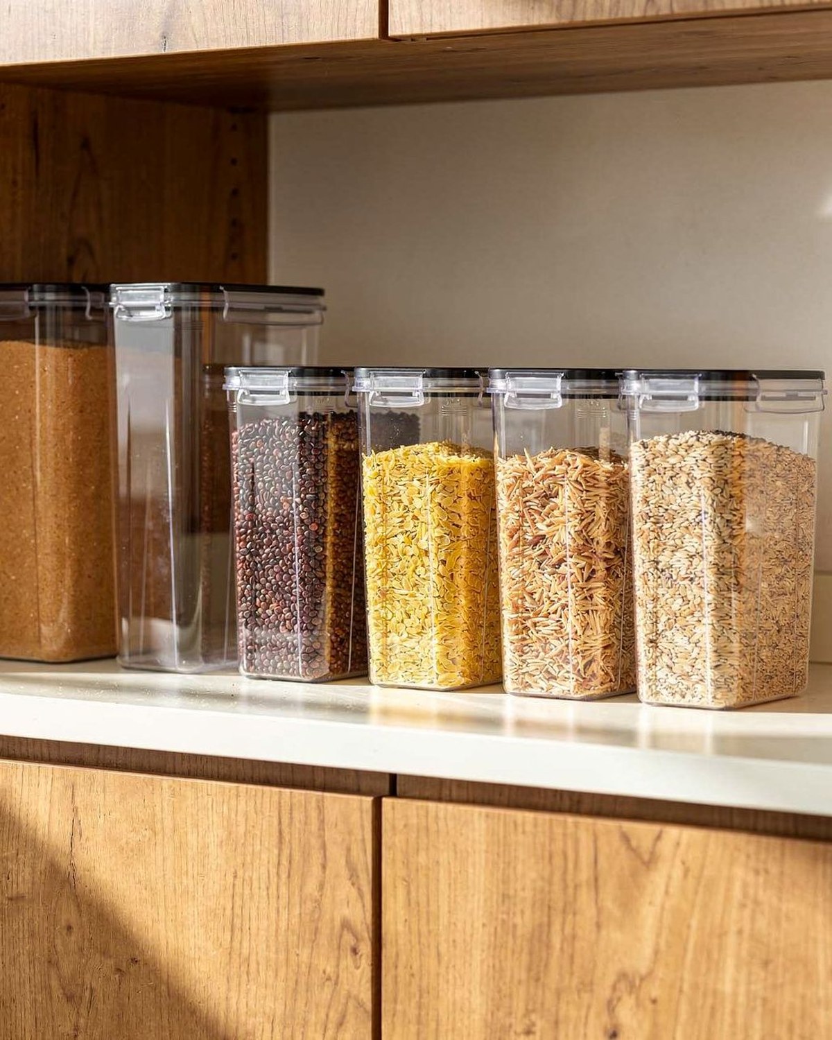 A matched 24-piece set of airtight pantry canisters with clear bodies, dark lids and chalk labels, lined up on a pantry shelf.