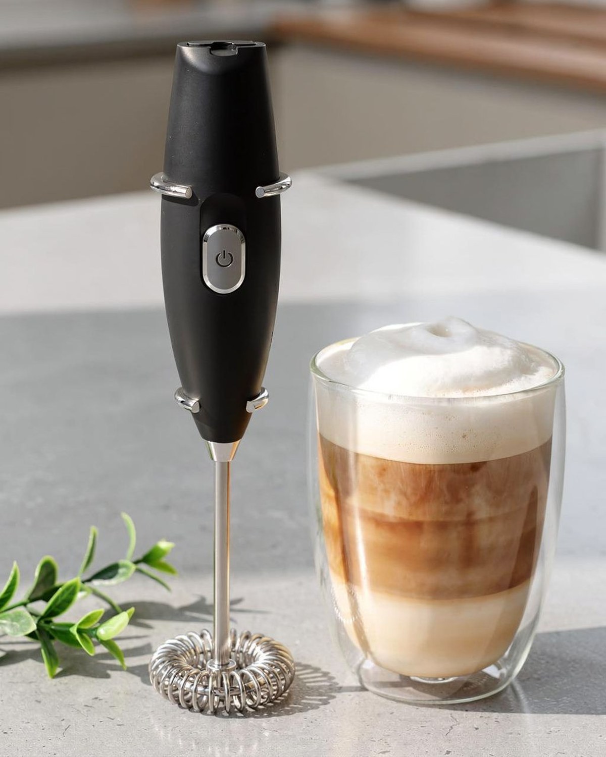 A slim black handheld battery frother wand beside a glass of layered iced coffee topped with foam.