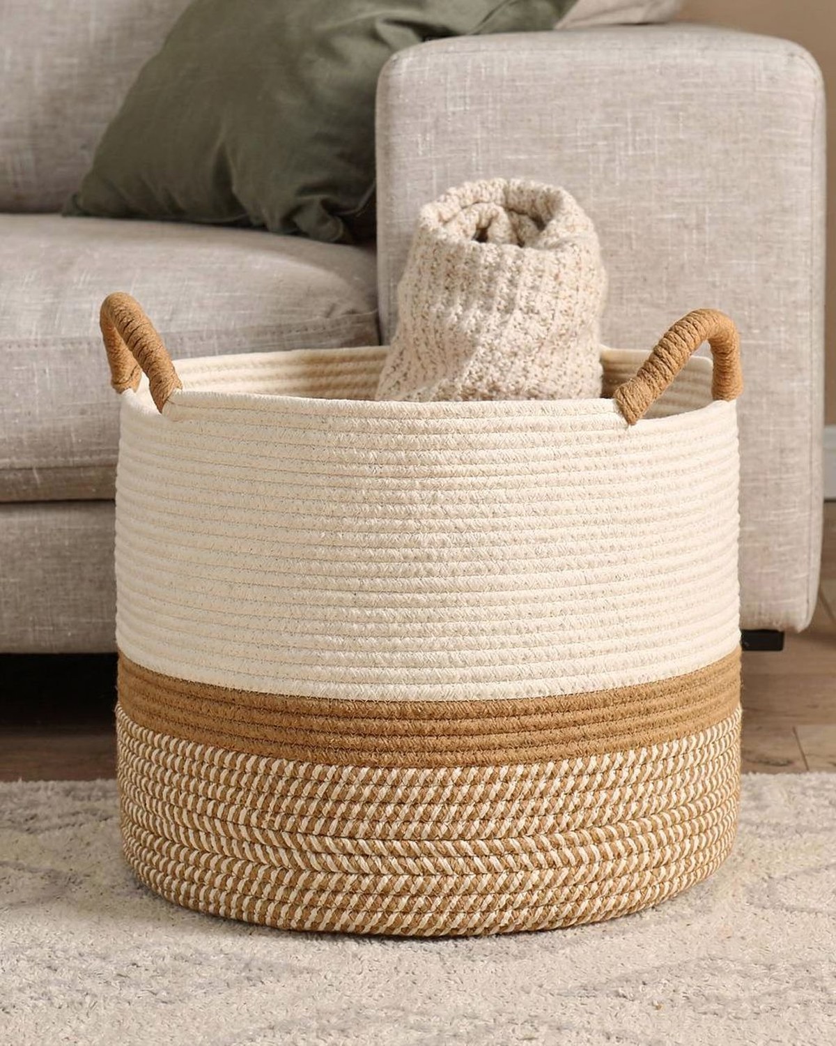 A large natural cotton-rope woven basket with handles, slouched open beside a sofa holding a rolled blanket.