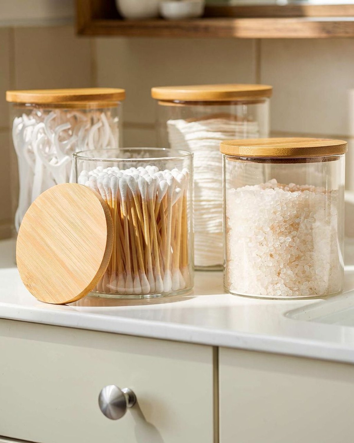 Four clear glass canisters with brushed-metal lids holding cotton balls, swabs, and bath salts, lined up on a vanity.