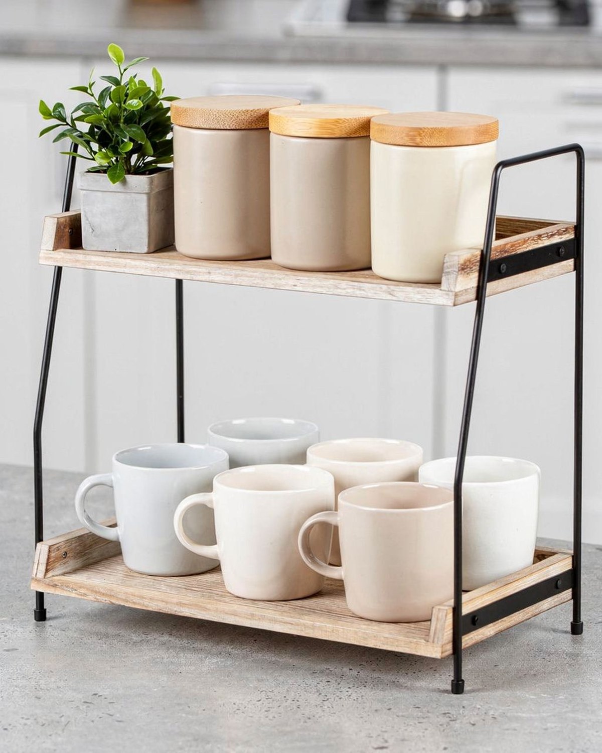 A two-tier wood-and-metal countertop shelf staged with mugs on the lower deck and canisters and a plant up top, forming a compact coffee corner.