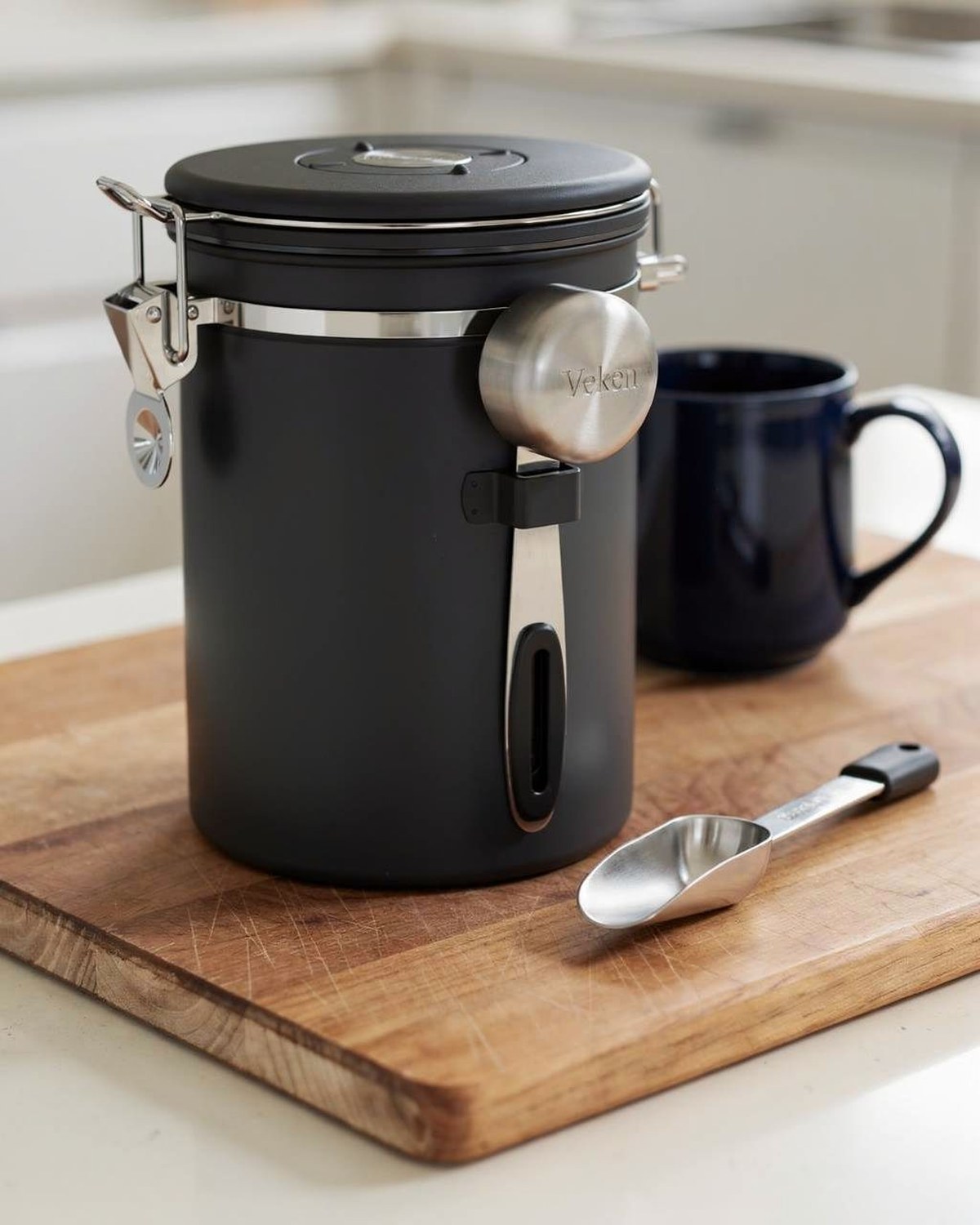 A matte stainless coffee canister with a front viewing window and a built-in scoop and date tracker on the airtight lid.
