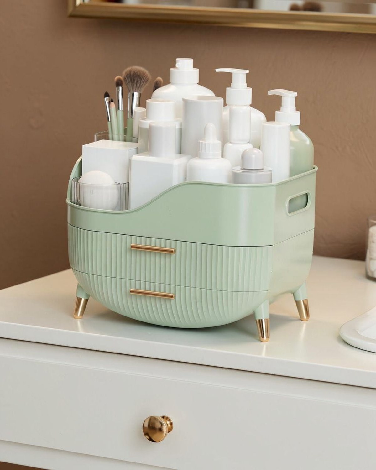 A sage-green countertop organizer with stacked clear pull-out drawers and an open top tray holding skincare bottles and brushes.