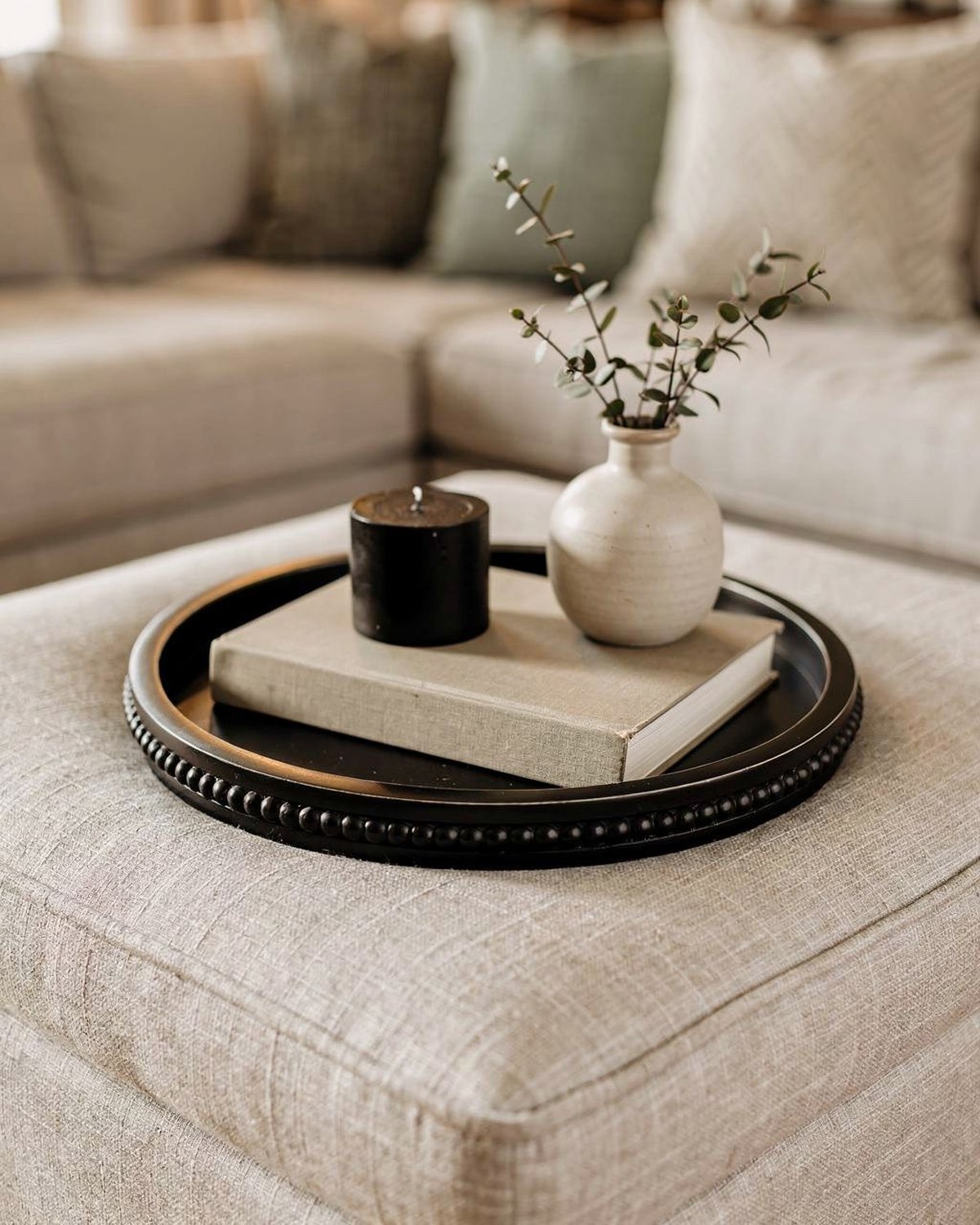 A round black-stained wood tray with low walls, styled on an ottoman with a candle, book and small vase.