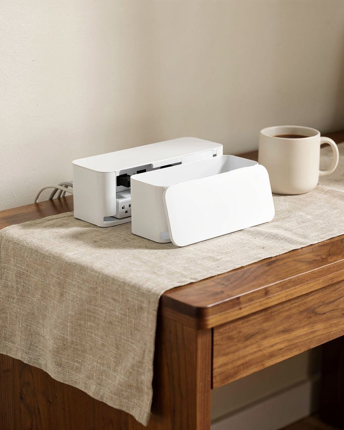 White cable management box on a desk edge concealing a power strip and routed charging cords